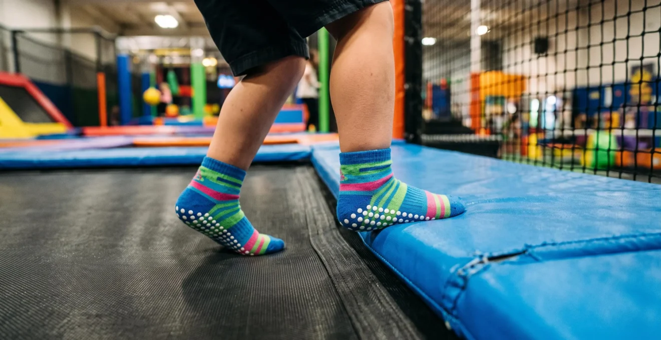 Gros plan sur les pieds d'un enfant portant des chaussettes antidérapantes colorées obligatoires, debout sur le bord d'un trampoline avec protection bleue